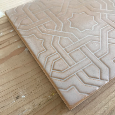 Azulejo cerâmico bege claro com padrão geométrico em relevo sobre superfície de madeira.