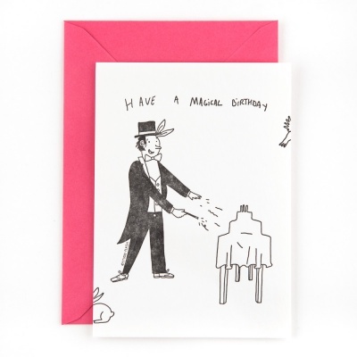 Cartão de aniversário com ilustração a preto de um mágico e texto 'HAVE A MAGICAL BIRTHDAY', e envelope rosa.
