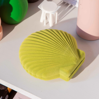Estojo verde em forma de concha com textura, sobre mesa branca junto a objetos decorativos