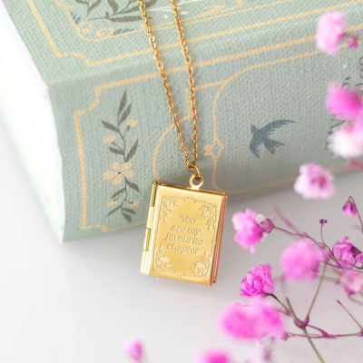 Colar dourado em forma de livro com texto gravado sobre um fundo com livro e flores cor-de-rosa