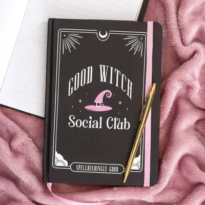 Caderno preto com texto GOOD WITCH Social Club e elástico rosa sobre tecido rosa com caneta dourada
