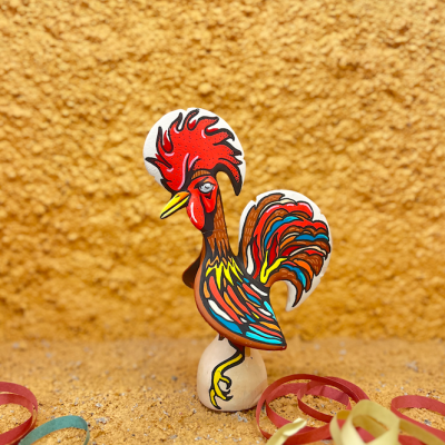 Miniatura de galo colorido em madeira com base branca sobre fundo amarelo texturizado e fitas coloridas