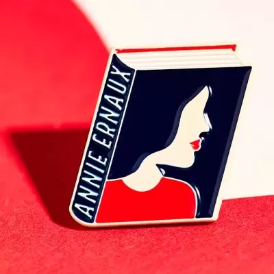 Pin em forma de livro com ilustração de perfil feminino e texto ANNIE ERNAUX