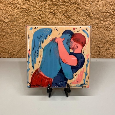 Ilustração de dois homens a abraçar-se, um azul com asas e outro ruivo, moldura em suporte preto.