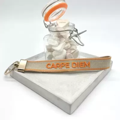 Pulseira cinza com texto laranja CARPE DIEM junto a frasco de vidro com tampa metálica