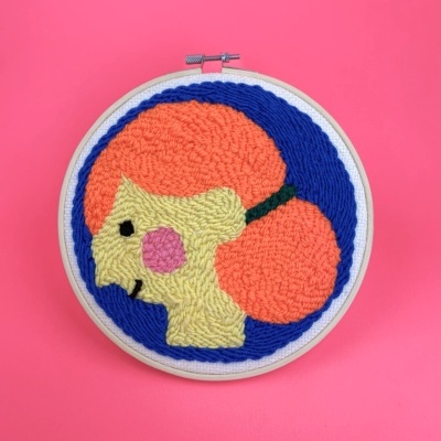 Aro de madeira com bordado colorido em forma de perfil feminino sobre fundo rosa