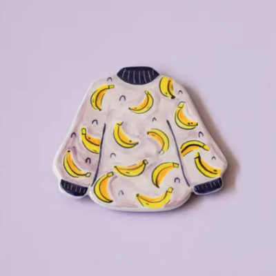 Prato branco formato camisola com estampa de bananas amarelas e detalhes azuis sobre fundo lilás.