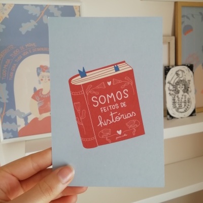 Cartão azul com ilustração de livro vermelho e texto 'SOMOS FEITOS DE histórias' segurado à frente de prateleira com quadros