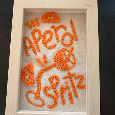 Quadro pequeno com bordado em lã laranja sobre tecido bege e moldura de madeira clara