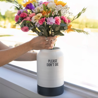 Vaso branco com base preta e texto PREASE DON'T DIE cheio de flores coloridas numa janela com mãos femininas