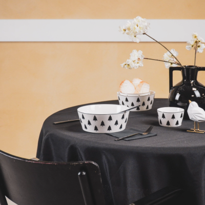 Mesa redonda com utensílios de mesa brancos com padrão de árvores pretas e jarra preta com flores, sobre toalha preta