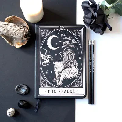 Caderno preto decorado com desenho branco de mulher lendo, lua e dragão, com lápis, vela e pedras ao redor