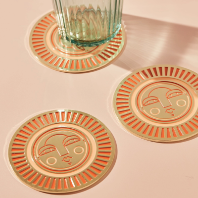 Três bases para copos com rosto estilizado em tons laranja e bege e um copo de vidro sobre uma mesa rosa