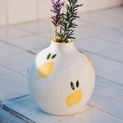 Vaso de cerâmica branco com estampas de limão, com plantas dentro, sobre azulejos.
