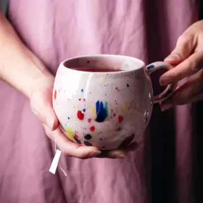 Caneca de cerâmica branca com manchas coloridas segurada por mãos, fundo desfocado e avental rosa-púrpura