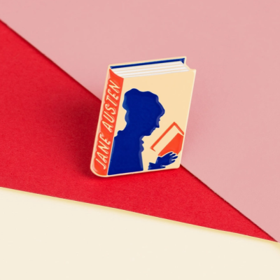 Broche esmaltado em forma de livro com silhueta de mulher a ler e texto JANE AUSTEN na lombada.