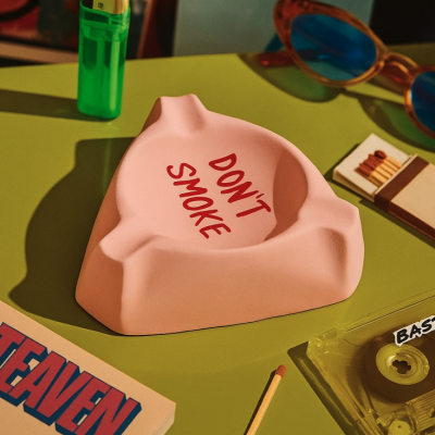 Cinzeiro cor-de-rosa com texto DONT SMOKE na superfície verde com isqueiro, fósforos, palito, óculos, cassete e livro.