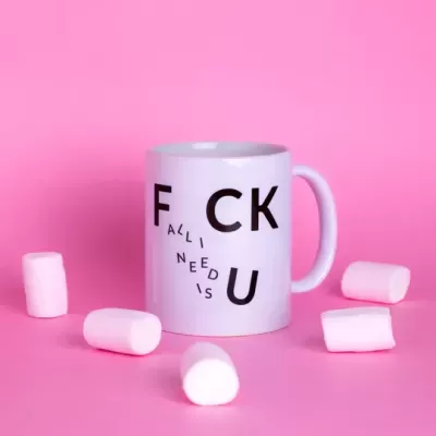 Caneca branca com texto preto 'F CK U' e 'ALL I NEED IS', rodeada por marshmallows brancos, fundo rosa