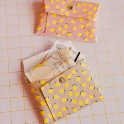 Duas carteiras pequenas com padrão de limões amarelos, uma aberta com notas dentro e outra fechada numa superfície quadriculada rosa