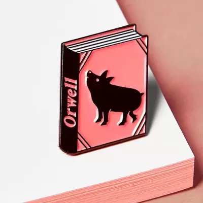 Pin em forma de livro cor de rosa com texto Orwell e porco preto