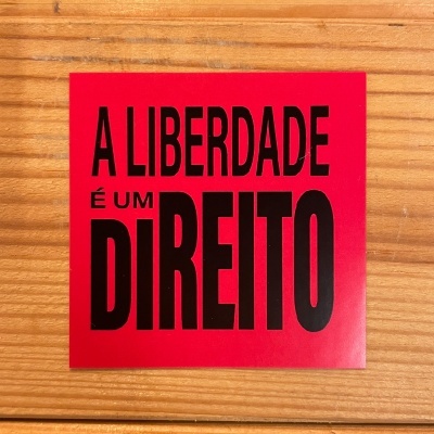 Autocolante vermelho com texto preto 'A LIBERDADE É UM DIREITO' em fundo de madeira