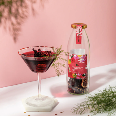 Copo de cocktail com bebida vermelha escura ao lado de frasco de vidro com tampa dourada e rótulo colorido