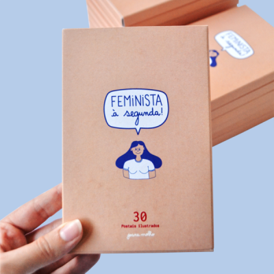 Caixa castanha com ilustração e texto Feminista à segunda, segurada por mão, fundo azul