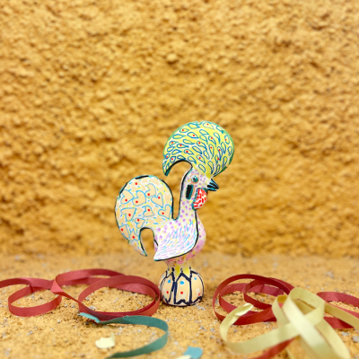 Estatueta colorida de galo com padrões e fitas coloridas em superfície castanha texturada.