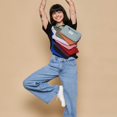 Mulher sorridente com cinco bolsas coloridas suspensas no peito em pose de yoga sobre fundo bege.