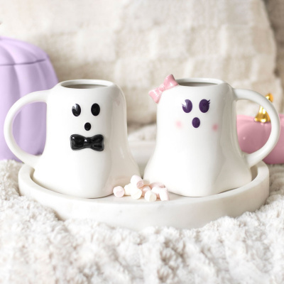 Duas canecas brancas em forma de fantasma com acessórios decorativos, numa bandeja branca com mini marshmallows.