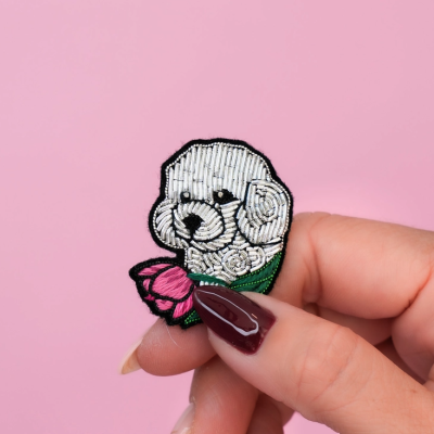 Broche bordado de cachorrinho branco com laço e flor segurado por mão com unhas vinho escuro