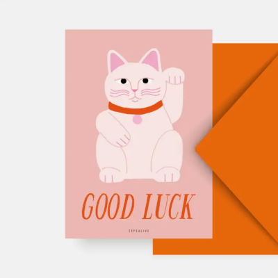 Cartão cor-de-rosa com gato branco e texto GOOD LUCK ao lado de envelope laranja