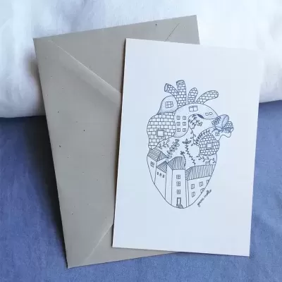 Cartão branco com desenho a preto de coração anatómico composto por edifícios, árvores e borboleta sobre envelope cinzento