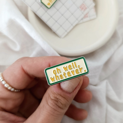Pin metálico retangular com texto oh well, whatever. em cores verde, branco e amarelo