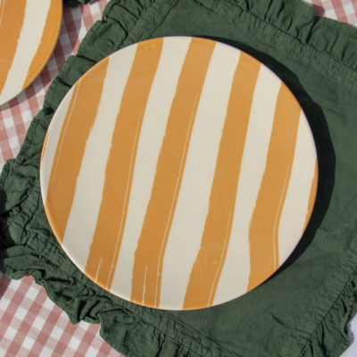 Prato de cerâmica com listras alaranjadas sobre fundo branco em toalha verde com babados e toalha xadrez rosa e branco