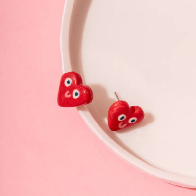 Brincos em forma de coração vermelho com olhos e boca sorridente sobre prato branco e fundo rosa