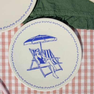 Prato de cerâmica branca com desenho azul de mulher na praia sobre toalha xadrez.