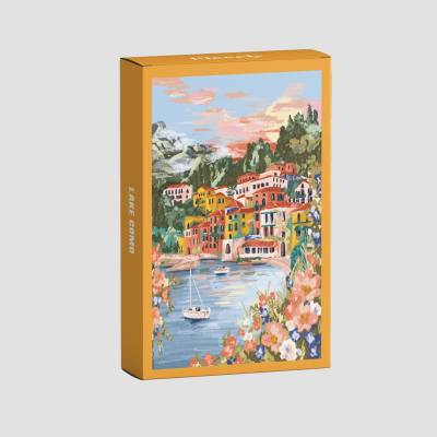 Caixa amarela com ilustração colorida de vila à beira de lago, barcos e flores, texto LAKE COMO na lateral.