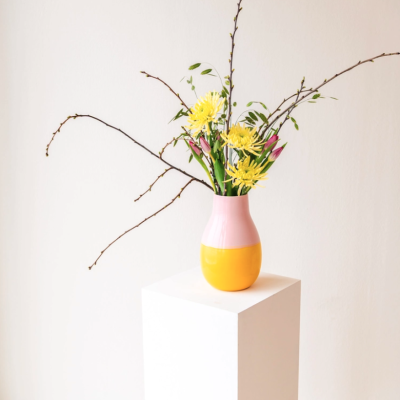 Vaso cerâmico rosa e amarelo com flores e ramos secos sobre pedestal branco