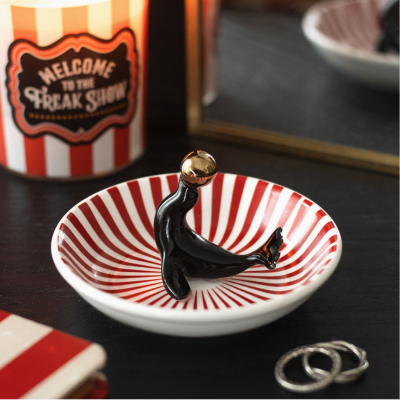 Prato decorativo vermelho e branco com figura de foca preta e dourada e caixa com texto Welcome to the Freak Show