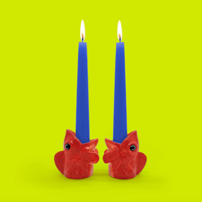 Dois castiçais vermelhos em forma de cabeça de peixe com velas azuis acesas em fundo amarelo.