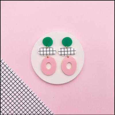 Brincos geométricos em verde, branco com padrão quadriculado preto e rosa, sobre fundo rosa
