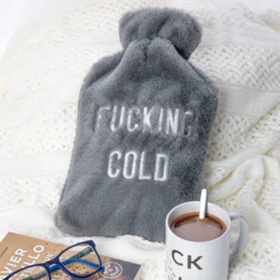 Frasco de água quente cinzento peludo com texto 'FUCKING COLD' numa manta branca, ao lado de uma caneca de bebida quente e óculos azuis.