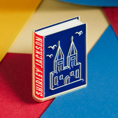 Pin em forma de livro azul com castelo e lombada vermelha com texto