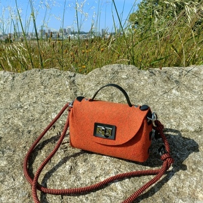 Mala pequena de tecido cor tijolo com alça trançada e fecho metálico sobre pedra