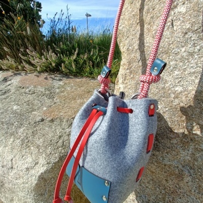Bolsa em tecido cinzento com detalhes em couro azul e vermelho pendurada em pedra ao ar livre
