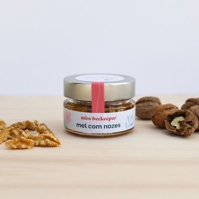 Frasco pequeno de mel com nozes com tampa prateada, nozes ao lado e superfície de madeira clara