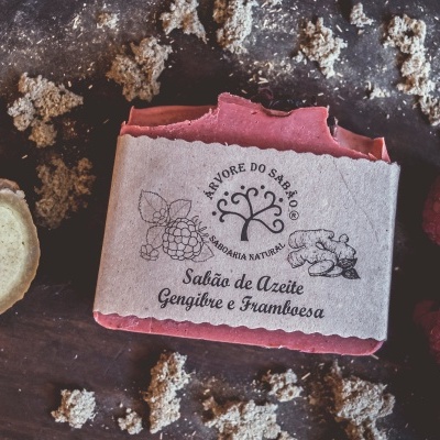 Sabão rosa de azeite gengibre e framboesa com embalagem de papel pardo sobre mesa escura com gengibre, framboesas e pedaços de areia.