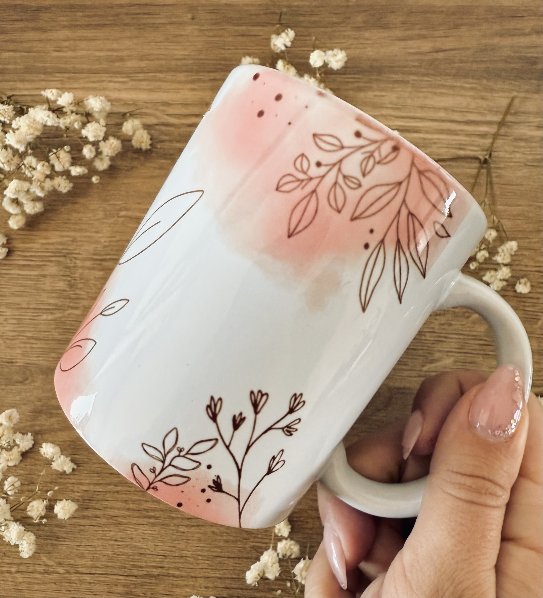 Caneca Flowers Caneca branca com padrão floral rosa e castanho, em mão sobre mesa de madeira.