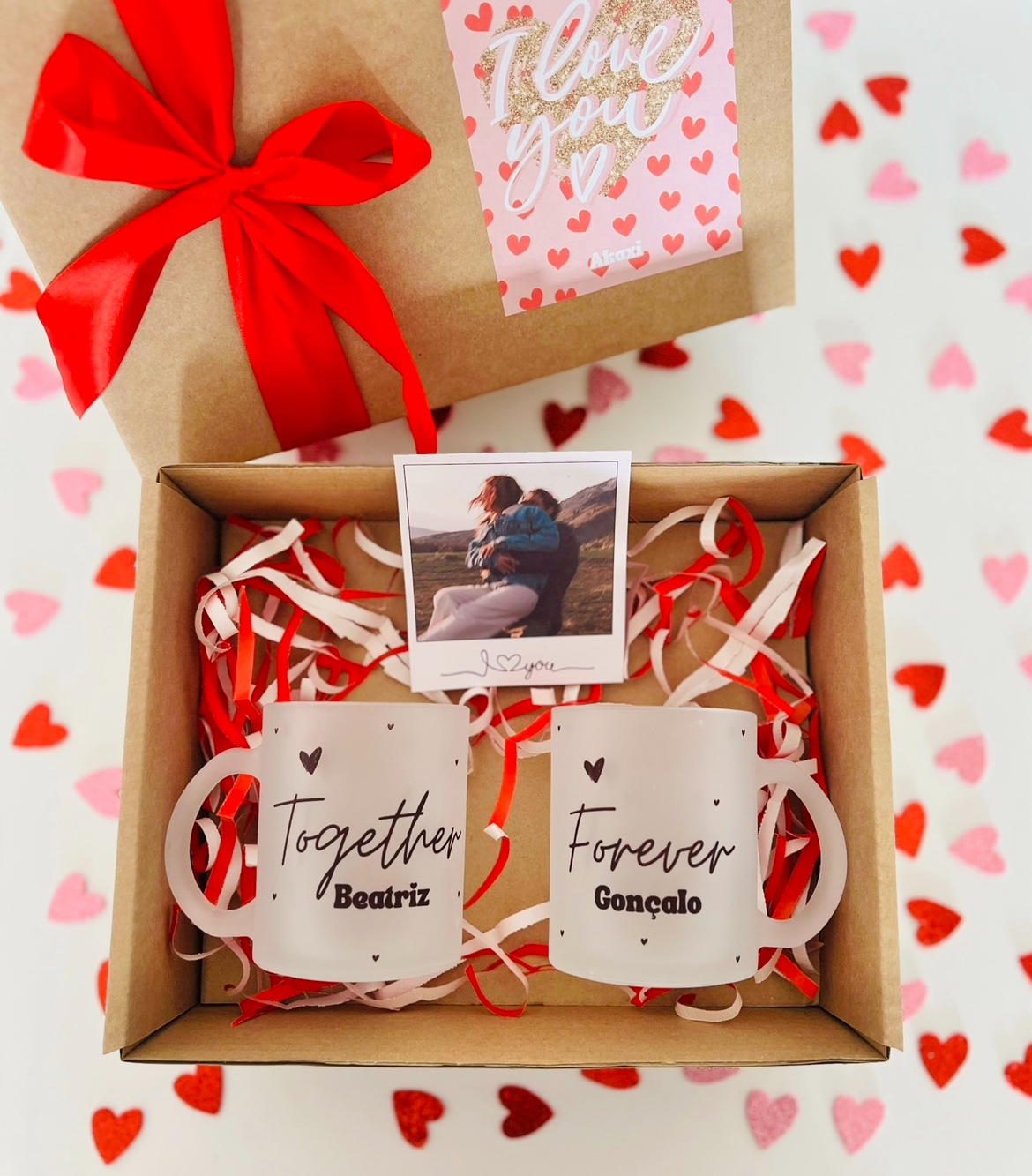 Box Canecas Together Forever Caixa de presente com dois copos personalizados com nomes e texto, com papel e foto, sobre superfície com padrão de corações